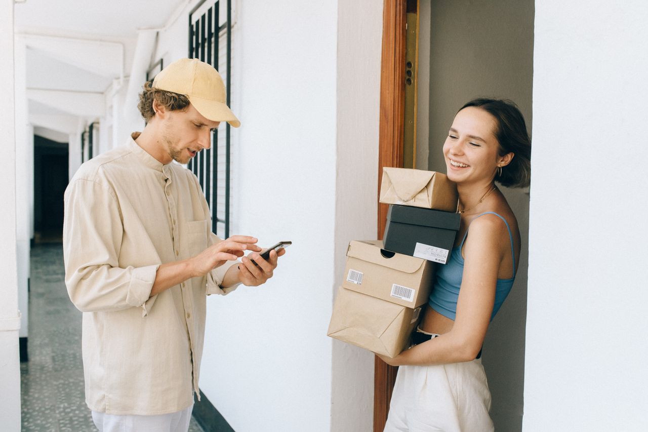 gallery-6 A cheerful woman receives a delivery from a person in a hallway, using a smartphone for online payment.